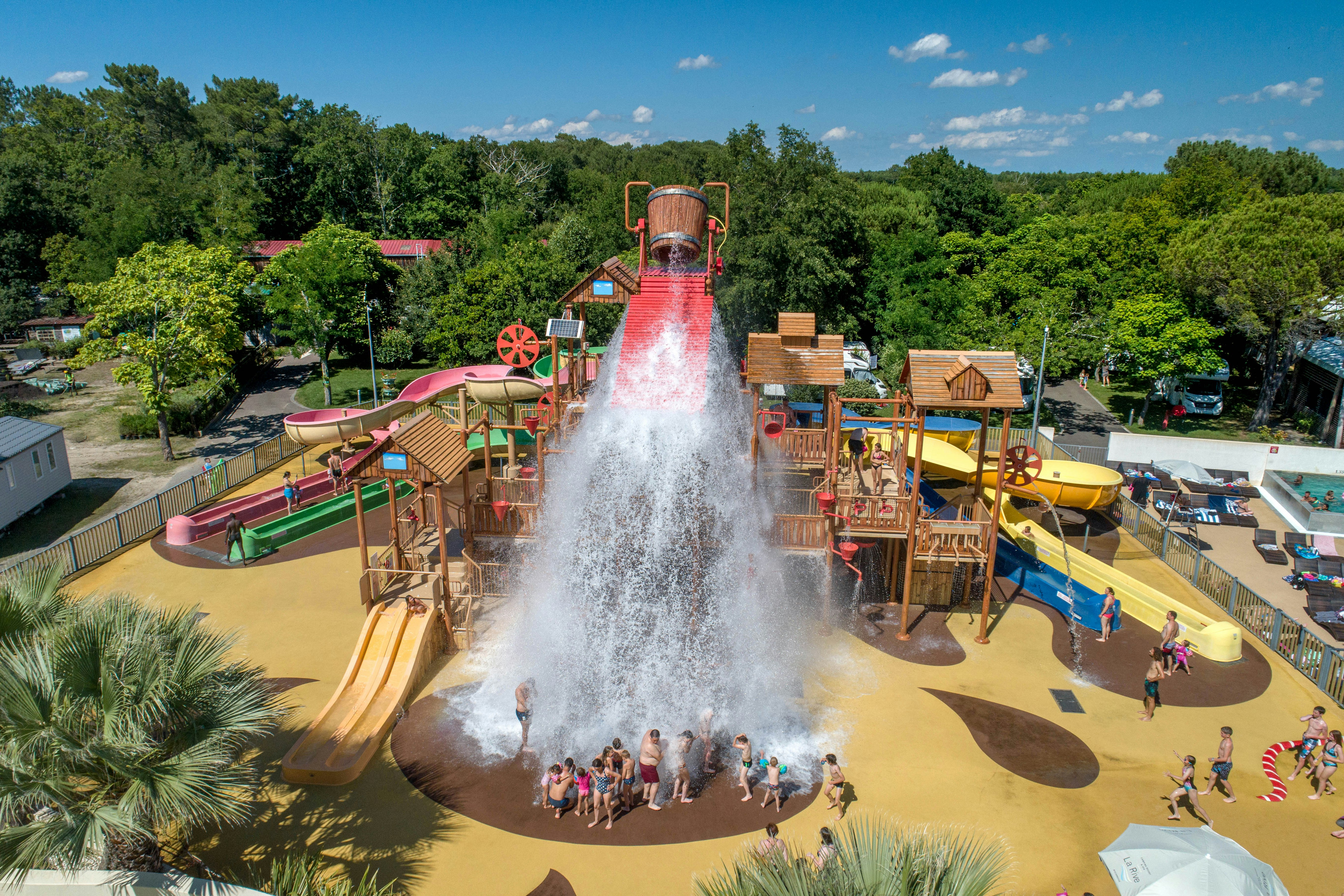 Wasserpark mit Sprühfontäne und Rutschen, Spielbereich mit Liegen und Bäumen im Hintergrund.