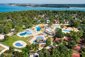 Luftaufnahme eines Campingplatzes mit mehreren Schwimmbecken, Sonnenschirmen, Liegen und Wegen im Grünen, am Seeufer.