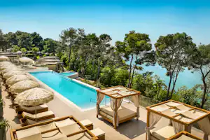 Blick von der Terrasse auf den Pool mit Sonnenliegen, Sonnenschirmen und Meerblick zwischen Bäumen im Campingbereich.