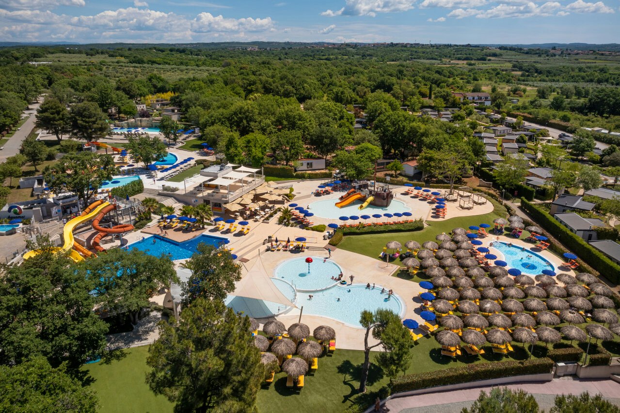 Luftaufnahme eines Campingplatzes mit mehreren Pools, Sonnenliegen, Wasserrutschen und einem großen Wasserbecken zwischen Bäumen.