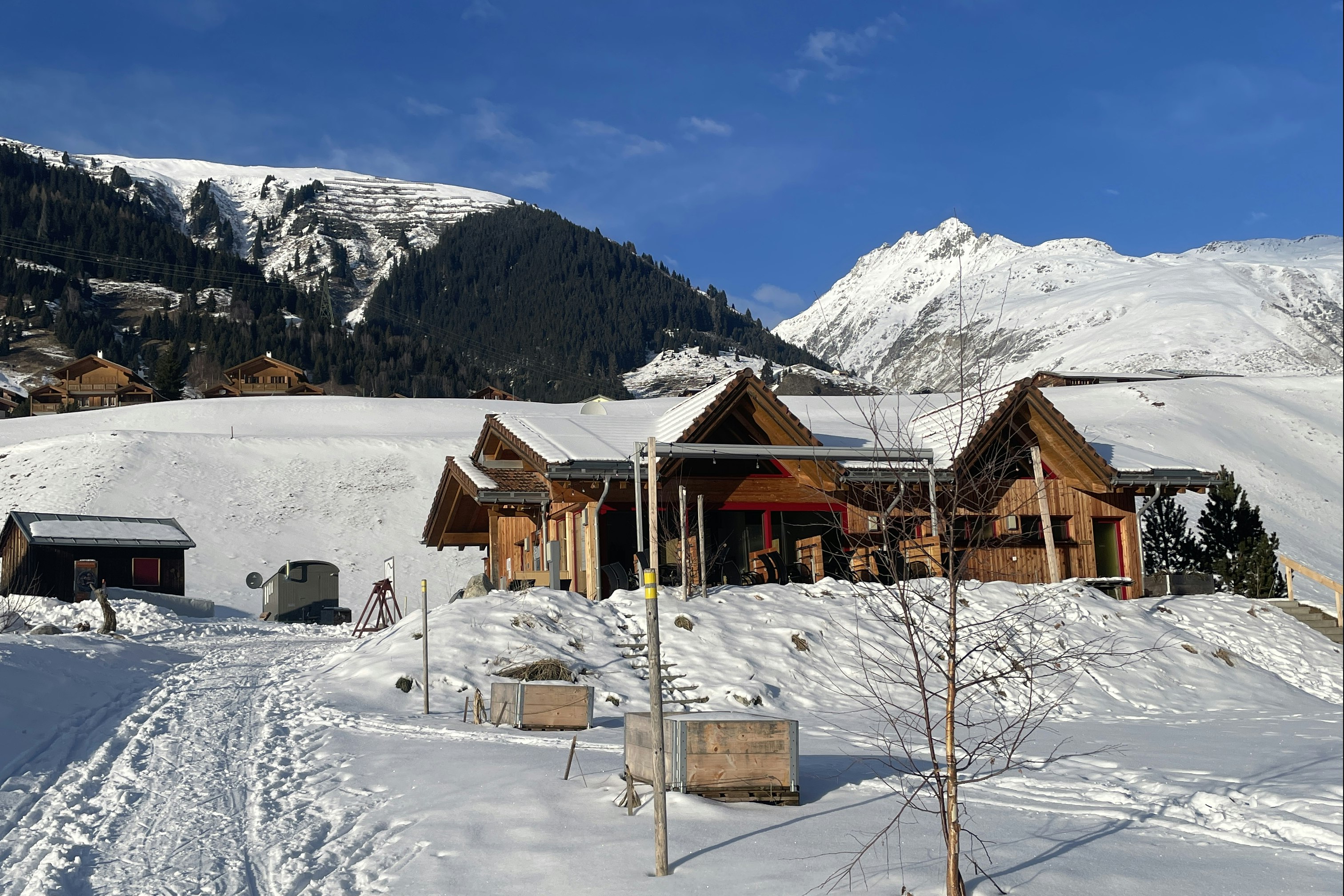 Winterliche Campinghütten mit Holzveranda und Sitzbank im Schnee, umgeben von Bergen und verschneiten Bäumen.