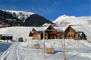 Winterliche Campinghütten mit Holzveranda und Sitzbank im Schnee, umgeben von Bergen und verschneiten Bäumen.