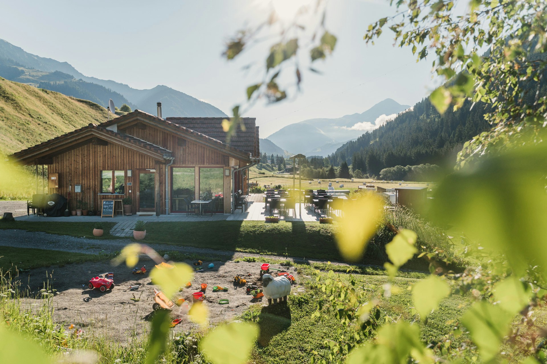 Campingplatz mit Holzgebäuden und Terrasse am Fluss, im Hintergrund Berge, davor Sitzgelegenheiten und Wege.