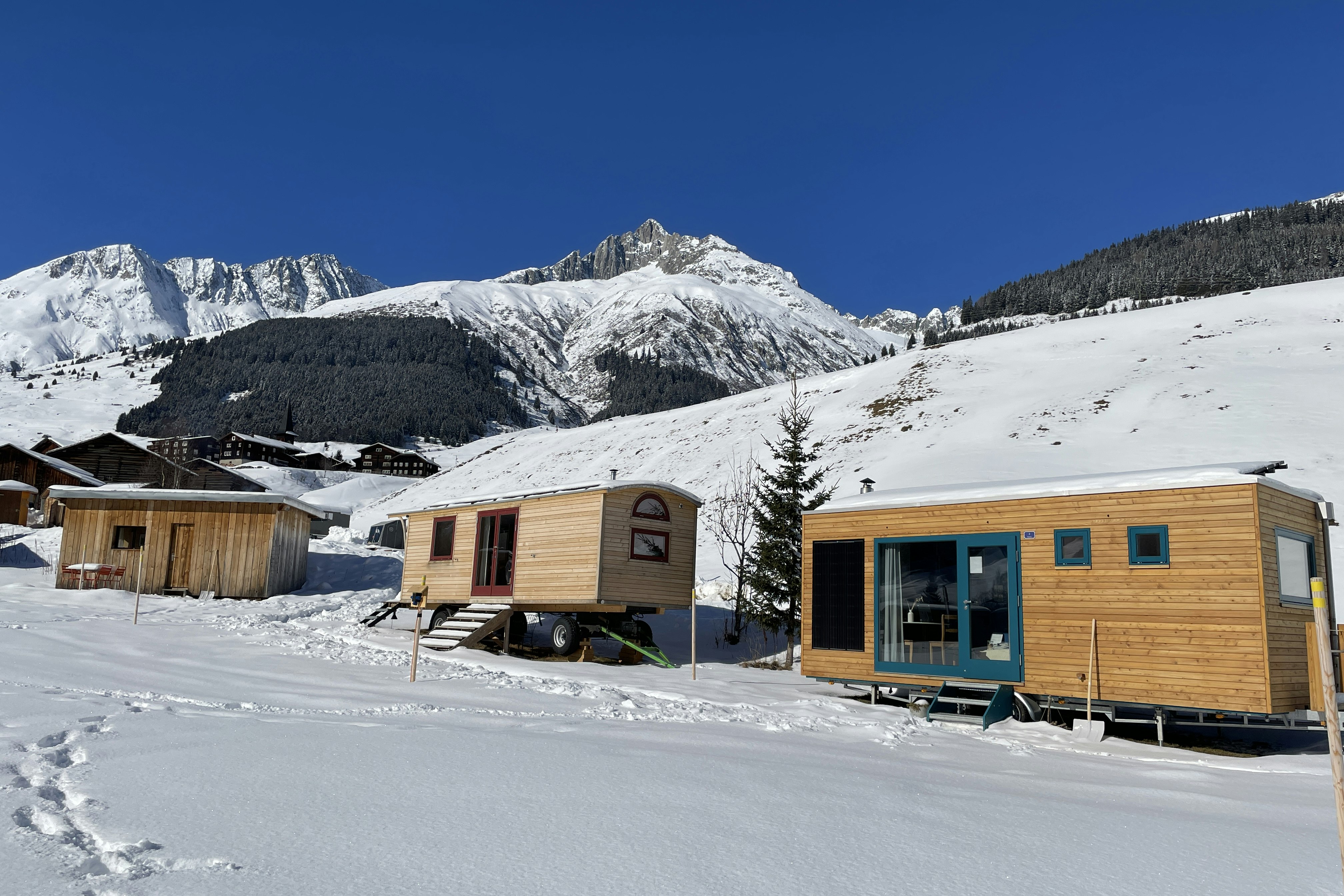 Holzhütten und ein Wohnwagen auf verschneitem Campingplatz, im Hintergrund Berge und klarer blauer Himmel.