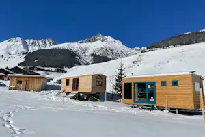 Holzhütten und ein Wohnwagen auf verschneitem Campingplatz, im Hintergrund Berge und klarer blauer Himmel.