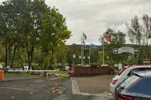 Eingang zum Campingplatz mit Schildern „Gasthof Rhönperle“, Flaggenmasten und Parkplatz am Rand, umgeben von Bäumen.