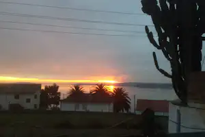 Sonnenuntergang über dem Meer, im Vordergrund ein Baum und Campinggebäude mit weißen Wänden und Palmen.
