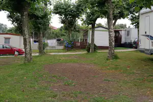 Stellplatz mit Wohnwagen und Vorzelt unter hohen Bäumen, Rasenfläche, Holzsichtschutz und Blumenbeet im Hintergrund.