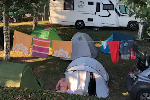 Campingzelt im Vordergrund, daneben mehrere Zelte und Wäscheleine; im Hintergrund steht ein Wohnmobil zwischen Bäumen.