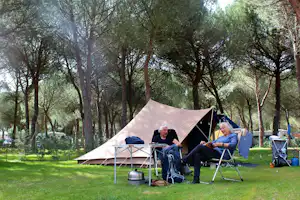 Camping im Grünen: Zelt auf dem Stellplatz, zwei Personen sitzen an Klappstühlen, daneben Tisch und Laterne im Schatten der Bäume.
