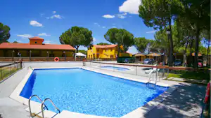 Großes Camping-Schwimmbecken mit Liegen und Sonnenschirmen, daneben Gebäude mit roten Dächern und Bäume im Hintergrund.