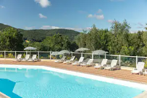 Camping-Pool mit blauem Wasser, Holzdeck und Liegen unter Sonnenschirmen, im Hintergrund grüne Bäume und Hügel.