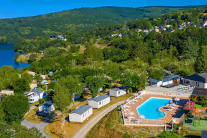 Blick auf Campingplatz mit Mobilheimen, Pool und Sonnenliegen, umgeben von Bäumen, Wegen und einem See im Hintergrund.