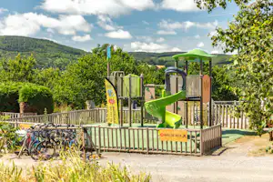 Spielplatz am Campingplatz mit grünem Rutschenturm, Zaun und Fahrrädern im Hintergrund, umgeben von Bäumen und Hügeln.