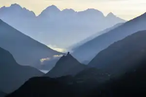 Bergpanorama mit tiefen Tälern und schroffen Gipfeln in den Alpen, im Hintergrund Dunstschichten und ein kleines Dorf.
