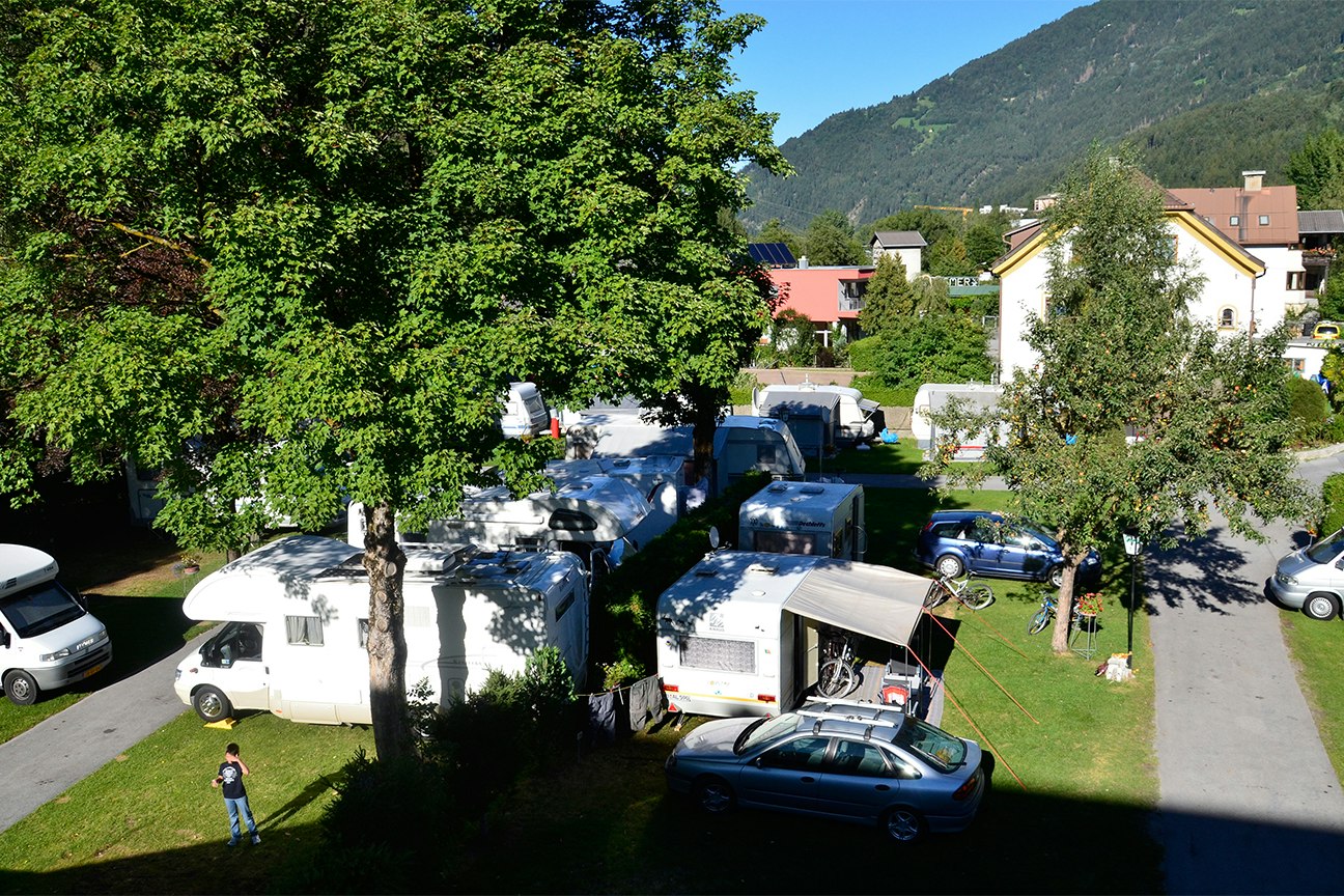 Campingplatz mit Wohnwagen und Autos auf dem Stellplatz, umgeben von Rasen, Bäumen und Häusern am Hang.