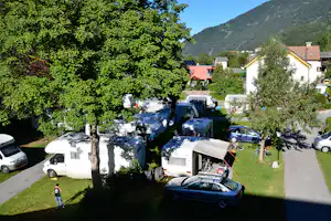 Campingplatz mit Wohnwagen und Autos auf dem Stellplatz, umgeben von Rasen, Bäumen und Häusern am Hang.