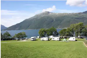 Wohnwagen auf einer grünen Wiese am See, im Hintergrund Berge und Bäume.