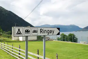 Hinweisschild „Ringøy“ am Campingplatz mit Wohnmobil- und Caravan-Symbolen, grüner Wiese, Zaun und Blick auf den See.