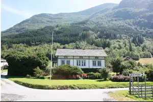 Weiße Ferienhaus-Vorderansicht mit gepflegtem Rasen, Hecke und Blumenbeeten, im Hintergrund bewaldete Berge und ein Weg.