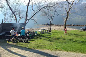 Grüner Rasen auf dem Campingplatz mit Grill, Sitzgruppe und Blick auf den See zwischen kahlen Bäumen.
