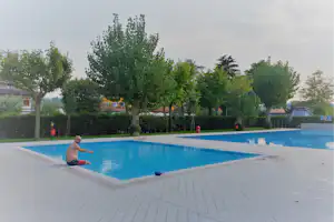 Schwimmbecken im Campingbereich mit blauem Wasser, gepflastertem Rand und Reihe von Bäumen im Hintergrund.