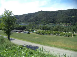 Campinganlage im grünen Tal mit Stellplätzen, Bäumen, Gebäuden und einem langen Gewächshausdach am Hang.