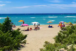 Sandstrand mit mehreren Sonnenschirmen und Liegestühlen, Urlauber am Wasser, im Vordergrund Büsche und Blick aufs Meer.