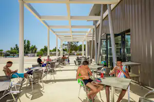 Überdachte Camping-Terrasse mit Liegen und Tischen, Kinder sitzen am Tisch, im Hintergrund Bäume und Sonnenschirme.