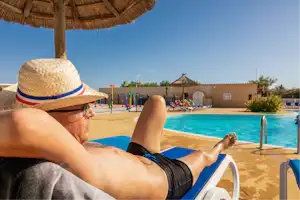 Mann mit Sonnenhut entspannt auf einer Liege am Pool, im Hintergrund Sonnenschirme, Liegestühle und ein Gebäude.