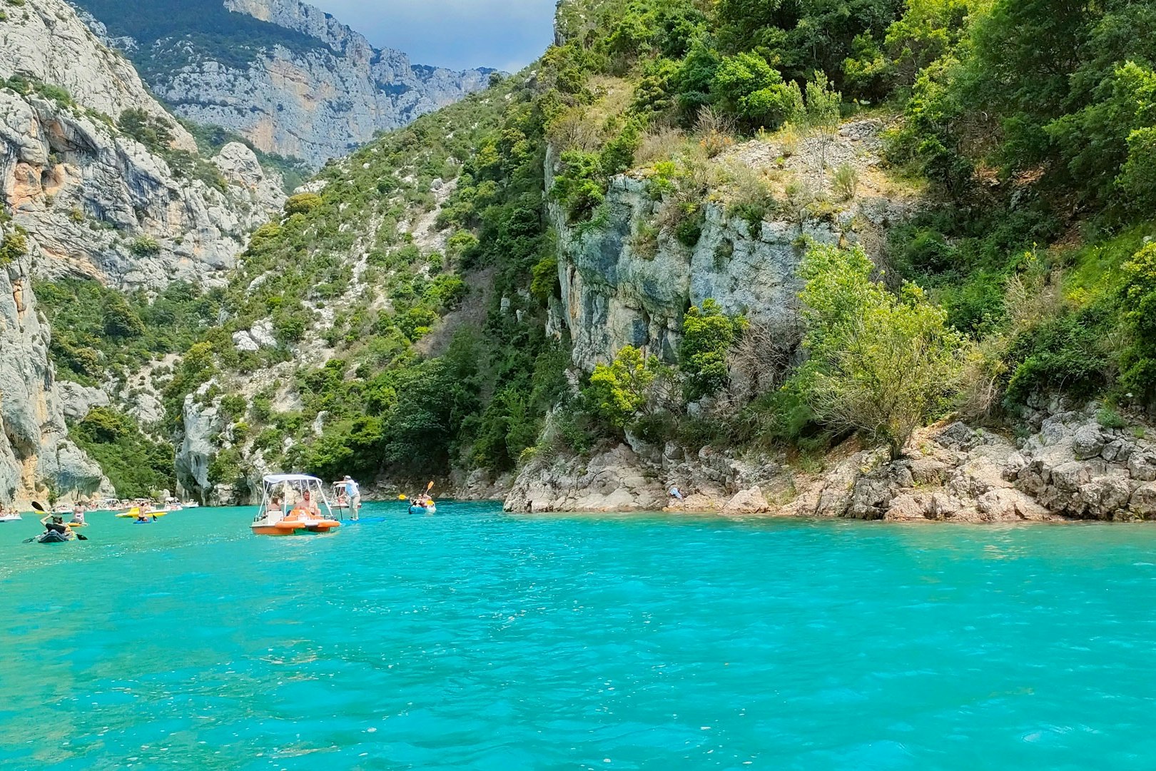 Türkisfarbenes Wasser vor steilen Felswänden und dichtem Wald, im Hintergrund Berge und ein kleines Boot auf dem See.