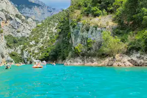 Türkisfarbenes Wasser vor steilen Felswänden und dichtem Wald, im Hintergrund Berge und ein kleines Boot auf dem See.
