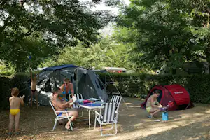 Campingplatz im Schatten: Zelte, Campingstühle und ein Tisch zwischen Bäumen, daneben ein rotes Zelt und ein Auto.