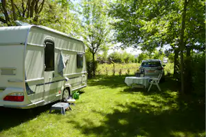 Wohnwagen auf einem grünen Campingplatz mit Picknicktisch und Gartenmöbeln, daneben ein Auto und Bäume im Hintergrund.