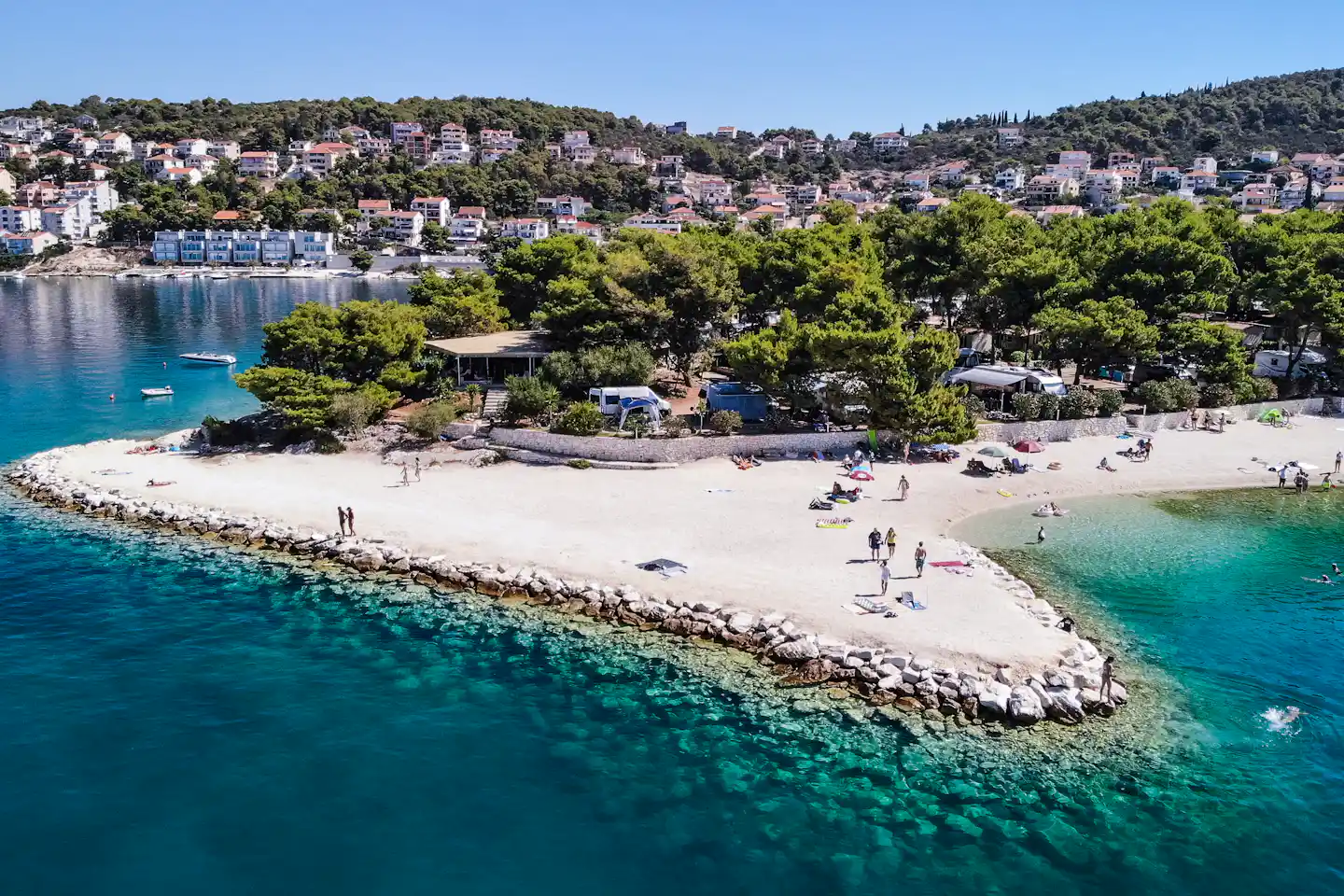 Badebucht mit Sandstrand und türkisfarbenem Meer, Wohnhäuser am Hang und Bäume am Ufer.