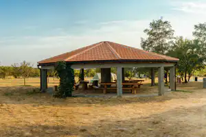 Campingunterstand mit Ziegeldach, Holztischen und Bänken, daneben Zelte im Gras und Bäume am Rand des Platzes.