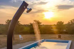 Außendusche am Campingplatz mit Poolrand, Blick in den Sonnenuntergang und Bäume im Hintergrund.