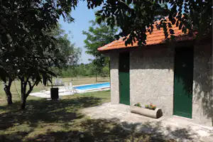 Steinwand mit grüner Tür und Dachziegeln, im Hintergrund Pool und Garten mit Bäumen und Liegestühlen.