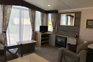 Wohnbereich in einem Mobilheim: Sofa, Holztisch, TV am Schrank und Kamin, große Fenster mit Vorhängen.
