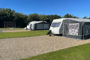 Zelte und Wohnwagen auf einem Kiesplatz, mit Rasenflächen und Hecken im Hintergrund.