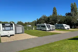 Wohnmobile auf Stellplätzen mit Betonweg und Rasenfläche, im Hintergrund Bäume und ein Wohnwagen unter klarem Himmel.