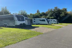 Wohnmobile auf einem Campingplatz, mit Kiesweg und Rasenfläche, im Hintergrund Bäume und weitere Fahrzeuge.