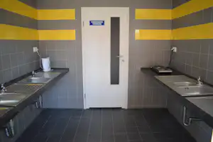 Gemeinschaftsküche mit zwei Spülbecken, Wasserkocher und Herdplatte auf Arbeitsplatten, Fliesenwände und Holztür im Hintergrund.