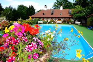 Blumenbeet im Vordergrund vor einem blauen Camping-Pool, dahinter ein weißes Gebäude mit Ziegeldach und Bänken.
