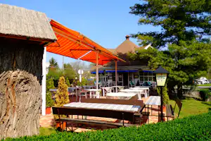 Camping-Terrasse mit roten Markisen, Tischen und Stühlen, im Hintergrund Gebäude und Bäume im Garten.