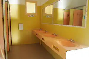 Camping-Badezimmer mit langen Waschbecken, Armaturen und Spiegeln, daneben Toilettenraum-Türen und Fliesenboden.