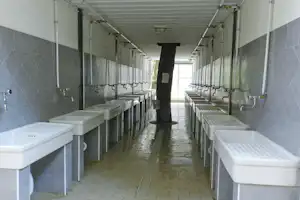 Waschraum mit mehreren Waschbecken in Reihen, grauen Fliesen und Handtuchhaltern, langer Flur mit Türen und Beleuchtung.