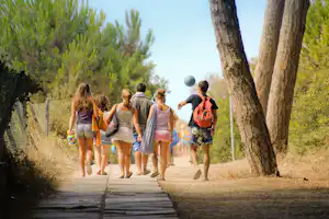 Familie geht auf einem Campingweg zwischen Bäumen, mit Rucksäcken und Kindern, im Hintergrund weitere Personen.