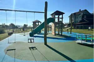 Spielplatz mit Rutsche, Schaukeln und Klettergerüst auf blauem Fallschutz, im Hintergrund Zäune und Wohnhäuser.