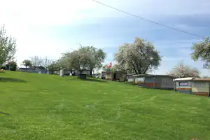 Grünes Campinggrundstück mit Rasenfläche, Wohnwagen und kleinen Gebäuden am Rand, Bäume und Wege im Hintergrund.